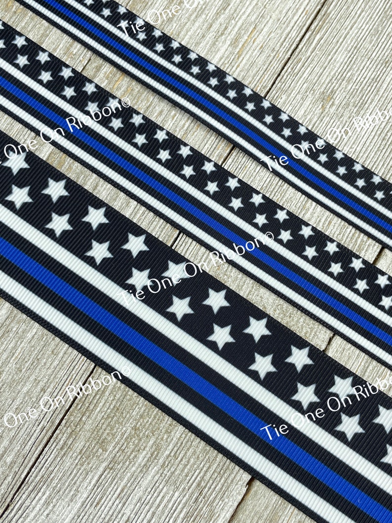May include: Three pieces of black and white ribbon with a thin blue stripe. The ribbon has white stars on a black background. The ribbon is for sale on Tie One On Ribbon.