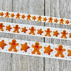 May include: Three white ribbons featuring a repeating pattern of gingerbread men and stars in orange and brown. The ribbons are arranged horizontally against a light wood grain background, ideal for festive crafts.