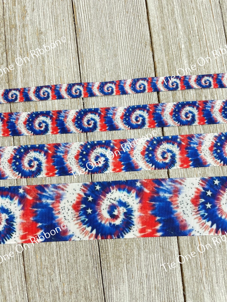 May include: Red, white, and blue tie dye ribbon with white stars. The ribbon is on a wooden surface.