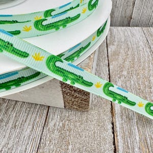 May include: A spool of light green ribbon featuring a repeating pattern of green alligators with blue accents and yellow floral elements. The ribbon is displayed on a white spool, with a wooden surface in the background.