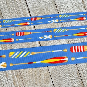 May include: Blue ribbon featuring a repeating pattern of oars in various colors, including red, yellow, and white. The oars have striped and solid designs. The ribbon is set against a weathered wood background.