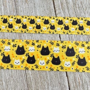 May include: Two ribbons with a repeating pattern of black and white cat faces on a yellow background. The ribbons are decorated with small leaves and dots. The background is a light wood grain.