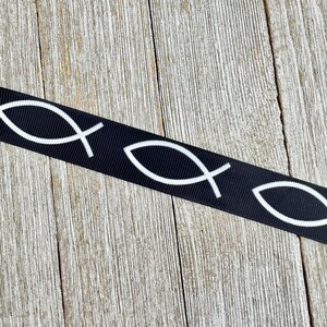 May include: Black ribbon featuring a repeating pattern of white Ichthys symbols, also known as the Jesus fish. The ribbon is displayed against a weathered, light brown wooden surface.