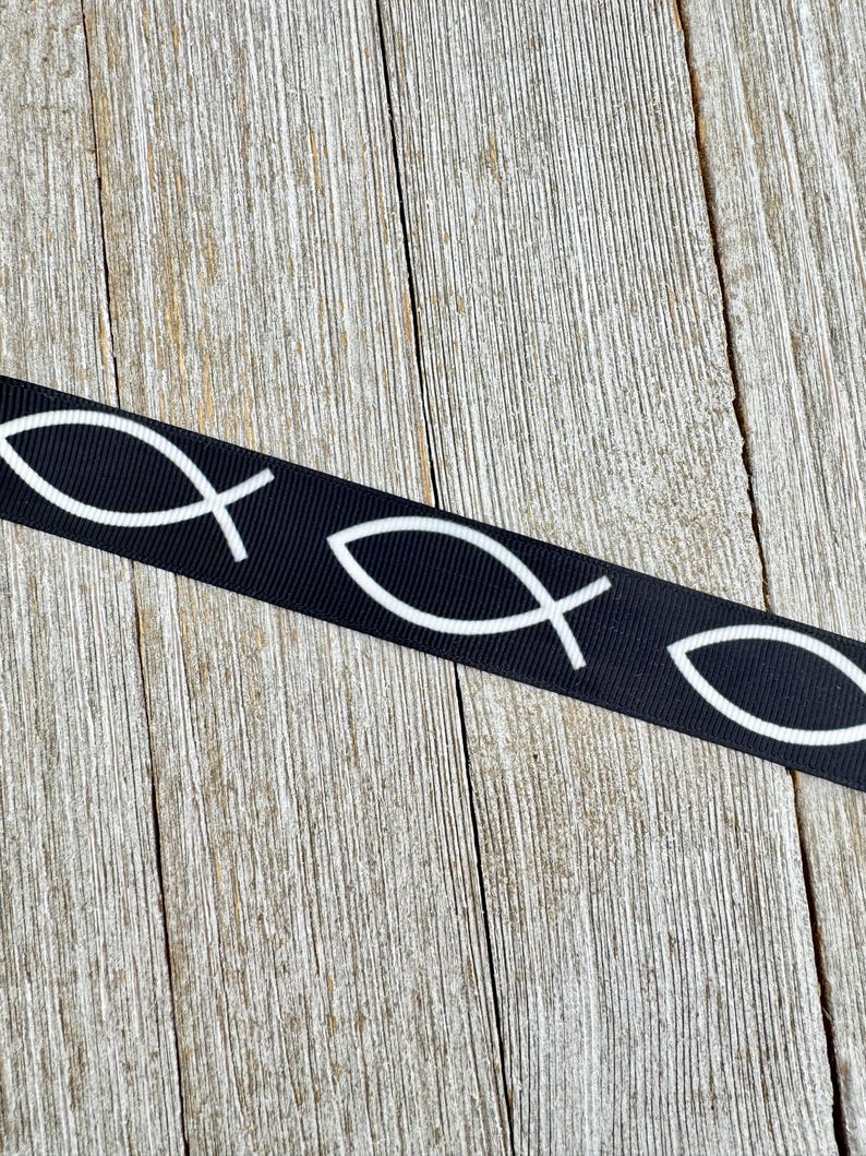 May include: Black ribbon featuring a repeating pattern of white Ichthys symbols, also known as the Jesus fish. The ribbon is displayed against a weathered, light brown wooden surface.