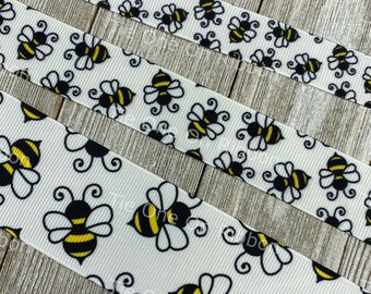 May include: White ribbon with a repeating pattern of black and yellow bees. The bees are outlined in black and have yellow stripes. The ribbon is on a wooden surface.