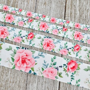 Könnte beinhalten: Weiße Bänder mit einem floralen Muster aus rosa Rosen, grünen Blättern und kleinen blauen Beeren. Die Bänder unterschiedlicher Breite sind auf einer Holzoberfläche angeordnet.