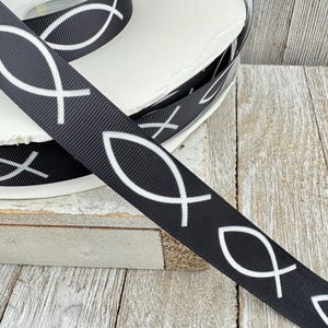 May include: Black ribbon with a white Ichthys symbol pattern. The ribbon is spooled on a white roll, with a close-up view of the detailed design. The ribbon is made of a durable material, suitable for various crafting projects.