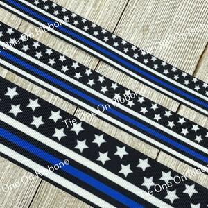 May include: Black and white ribbon with a thin blue stripe, featuring white stars on a black background. The ribbon has the text "Tie One On Ribbon" printed on it.