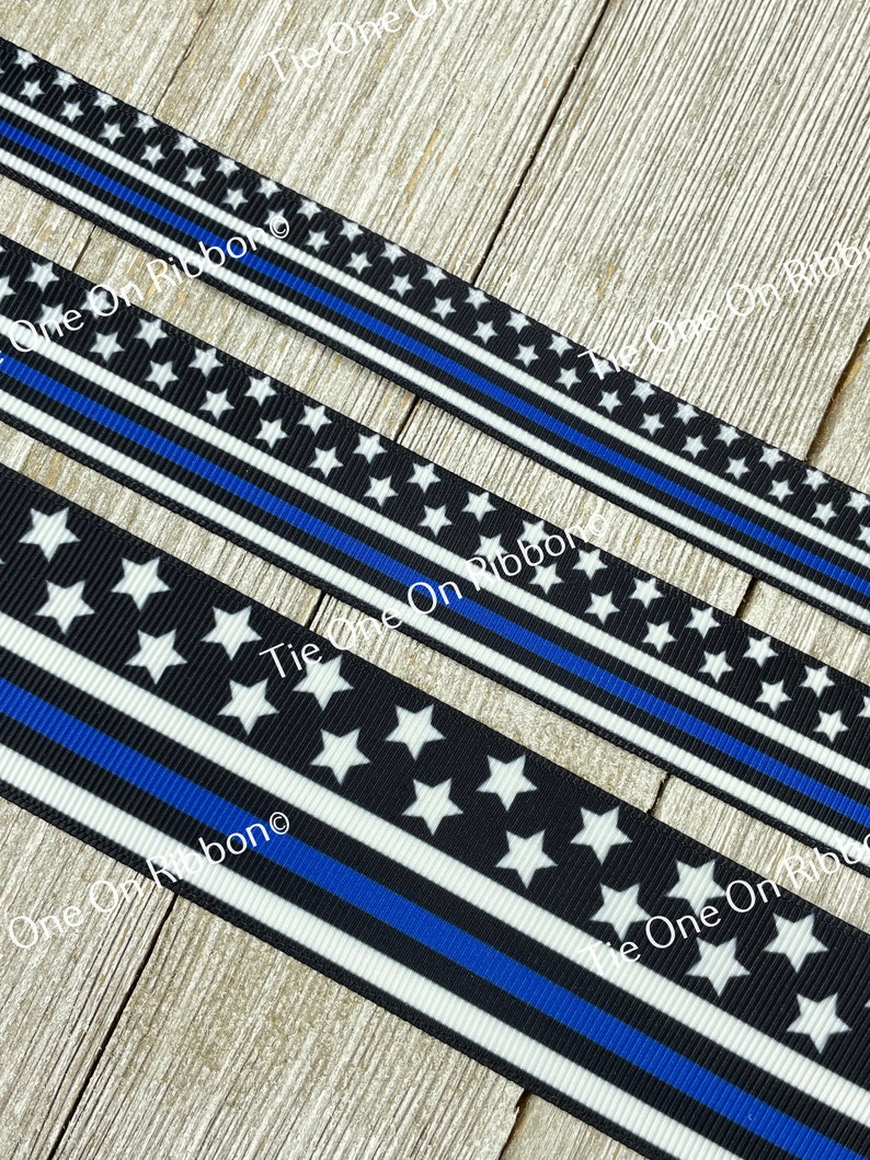 May include: Black and white ribbon with a thin blue stripe, featuring white stars on a black background. The ribbon has the text "Tie One On Ribbon" printed on it.
