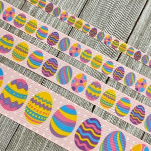May include: Pink ribbon with white polka dots and colorful Easter egg designs. The eggs feature various patterns and colors, including yellow, blue, purple, and pink. The ribbon is ideal for crafting and decorating.