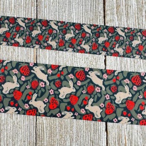 May include: Two strips of fabric with a repeating pattern of white rabbits, red strawberries, and pink flowers on a dark green background. The fabric is placed on a light wood surface.