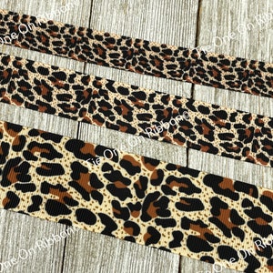 May include: Close-up of a brown, black, and cream leopard print ribbon. The ribbon is folded in thirds, with the top layer slightly offset from the bottom two layers.