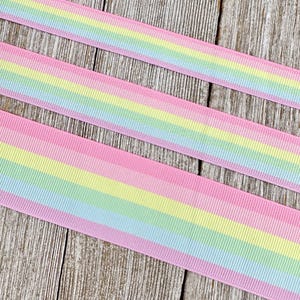 Puede incluir: Tres piezas de cinta rosa con rayas de arcoíris. La cinta está sobre una superficie de madera.