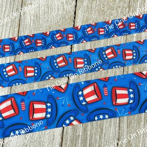 May include: Blue and white ribbon with a repeating pattern of red, white, and blue top hats with stars and fireworks. The ribbon is on a wooden surface.
