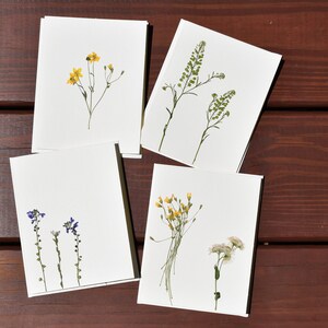 Puede incluir: Cuatro tarjetas blancas con flores prensadas. Las tarjetas presentan varias flores secas en amarillo, verde y morado. Las tarjetas están dispuestas sobre una superficie de madera marrón oscuro.