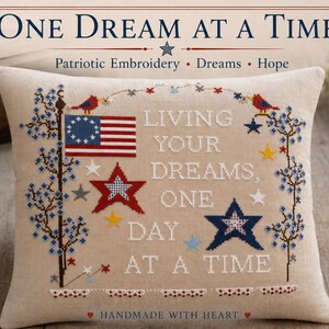 Può includere: Un cuscino quadrato beige con ricamo a punto croce che riporta il testo 'LIVING YOUR DREAMS, ONE DAY AT A TIME'. Il design include una bandiera americana rossa, bianca e blu, stelle rosse e blu, piccoli uccelli e tralci floreali blu.