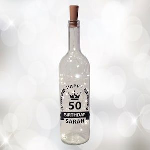 Könnte beinhalten: Eine klare Glasflasche mit einem Korkverschluss und einem schwarz-weißen Aufkleber mit der Aufschrift "Happy 50th Birthday Sarah" mit einem Kronen- und Lorbeerkranz-Design. Die Flasche ist mit weißen Lichterketten gefüllt.