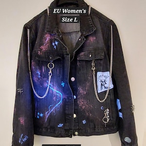 Puede incluir: Chaqueta vaquera negra con pintura reactiva a los rayos UV y detalles de cadena. Cuenta con un parche de ciervo y varios diseños pintados. Incluye el texto "EU Women's Size L" y "with UV light".