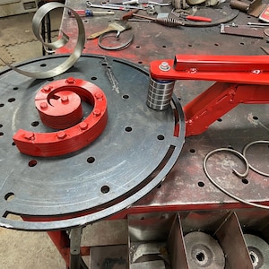 May include: A metalworking shop scene featuring a red metal spiral bending tool and a partially formed metal spiral. The tool is mounted on a red frame, with a circular metal plate. Various tools are scattered around.