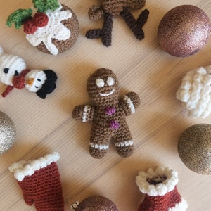 Pode incluir: Uma coleção de enfeites de Natal feitos à mão em crochê, incluindo um homem de gengibre, um boneco de neve, uma rena, um pudim e uma luva. Os enfeites são decorados com fio branco, marrom, vermelho e verde.