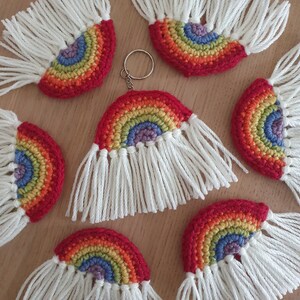 May include: A set of seven rainbow crocheted keyrings with white fringe. Each keyring features a rainbow with red, orange, yellow, green, blue, indigo, and violet colours. The keyrings are handmade and have a unique, whimsical design.