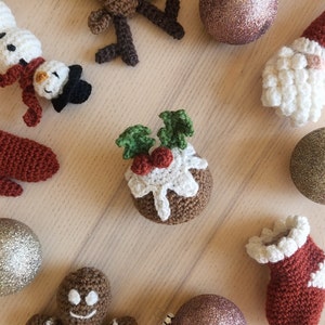 May include: A collection of crocheted Christmas ornaments, including a snowman, a reindeer, a stocking, a gingerbread person, a Santa Claus, and a Christmas pudding. The ornaments are arranged on a wooden surface.