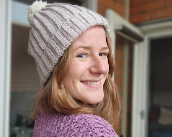 May include: A woman wearing a light pink crocheted beanie with a white pom pom. The beanie has a ribbed texture. She is wearing a purple sweater.