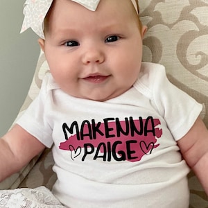May include: A baby girl wearing a white onesie with the text "Makenna Paige" in black lettering and a pink heart on each side of the text. The onesie has a pink watercolor-style background. The baby is wearing a white and gray bow headband and a pink and white tulle skirt.