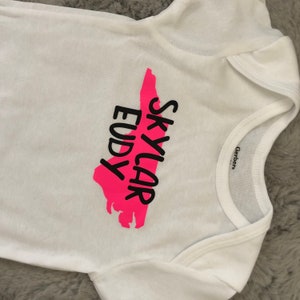 May include: White baby onesie with a pink outline of the state of North Carolina and the text "Skylar Eudy" in black.
