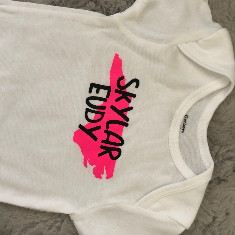 May include: White baby onesie with a pink outline of the state of North Carolina and the text "Skylar Eudy" in black.