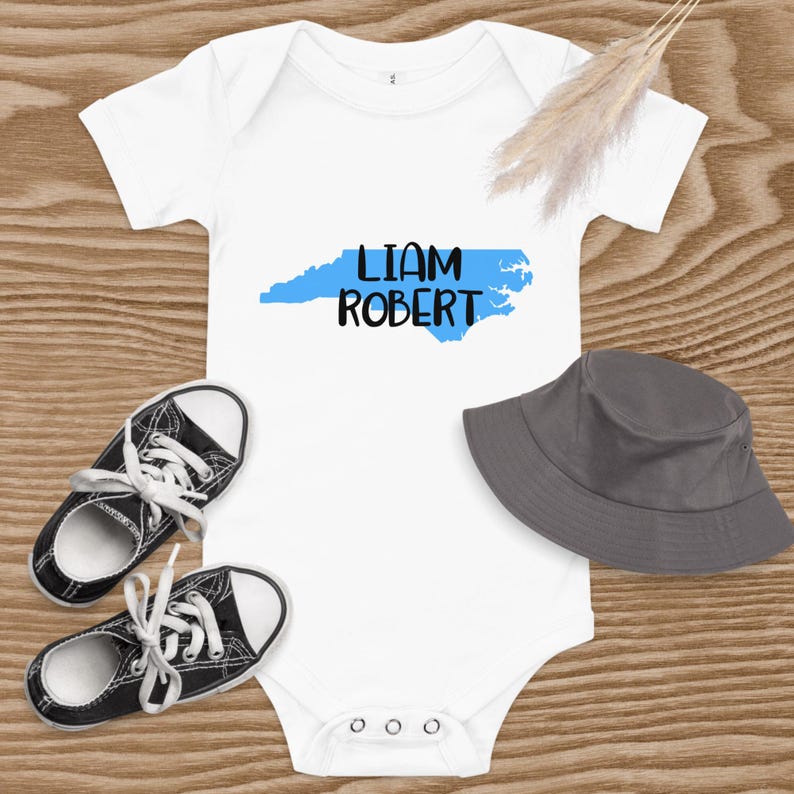 May include: White baby onesie with a blue outline of the state of North Carolina and the text "Liam Robert" printed in black.