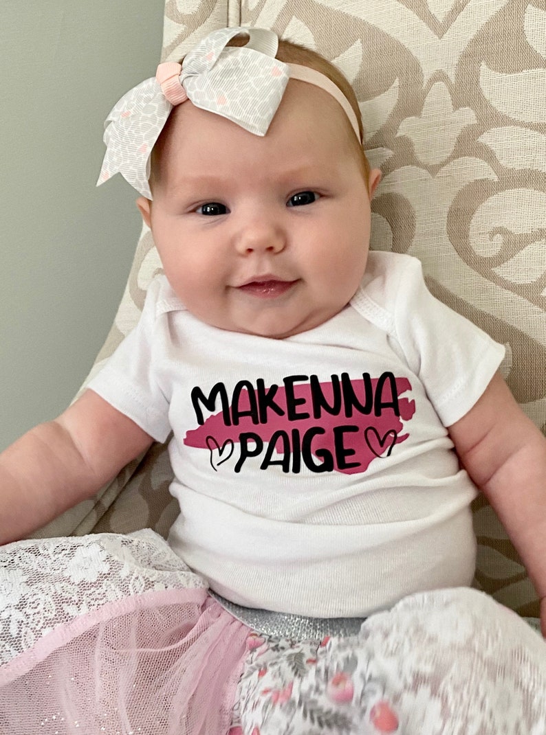 May include: A baby girl wearing a white onesie with the text "Makenna Paige" in black lettering and a pink heart on each side of the text. The onesie has a pink watercolor-style background. The baby is wearing a white and gray bow headband and a pink and white tulle skirt.
