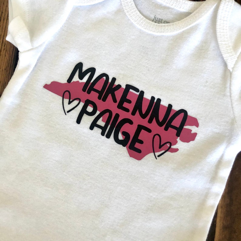 May include: White baby onesie with a pink outline of the state of North Carolina and the text "Makenna Paige" in black with two hearts.