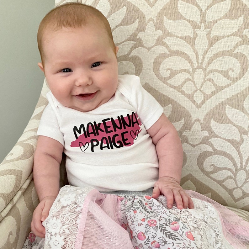 May include: A baby girl wearing a white onesie with the text "Makenna Paige" in black lettering and a pink heart on each side of the text. She is wearing a pink and white floral skirt with lace trim.