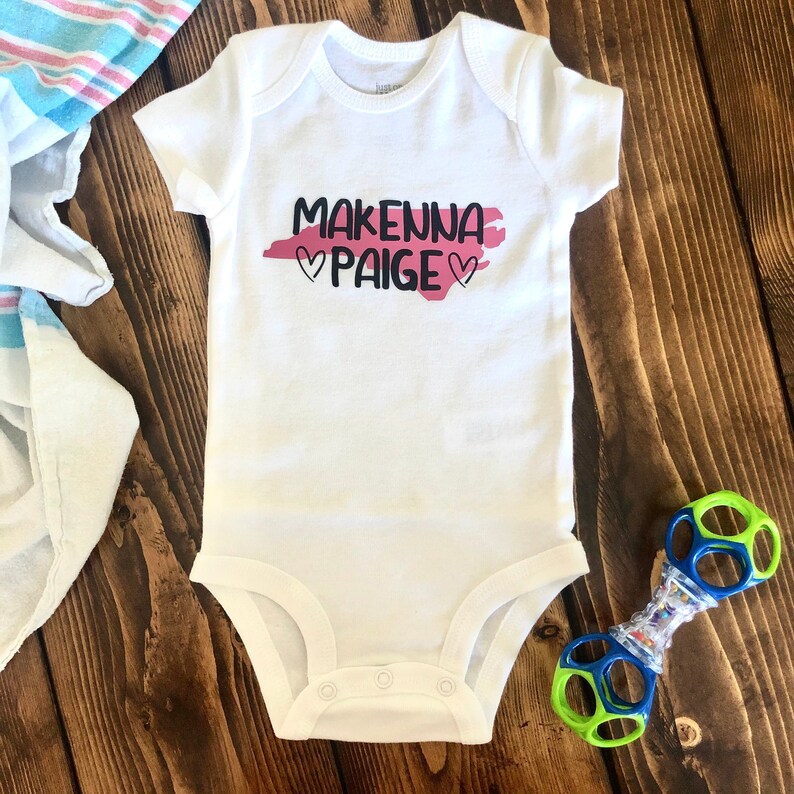 May include: White baby onesie with a pink and black design. The design features the names "Makenna Paige" with hearts on either side.