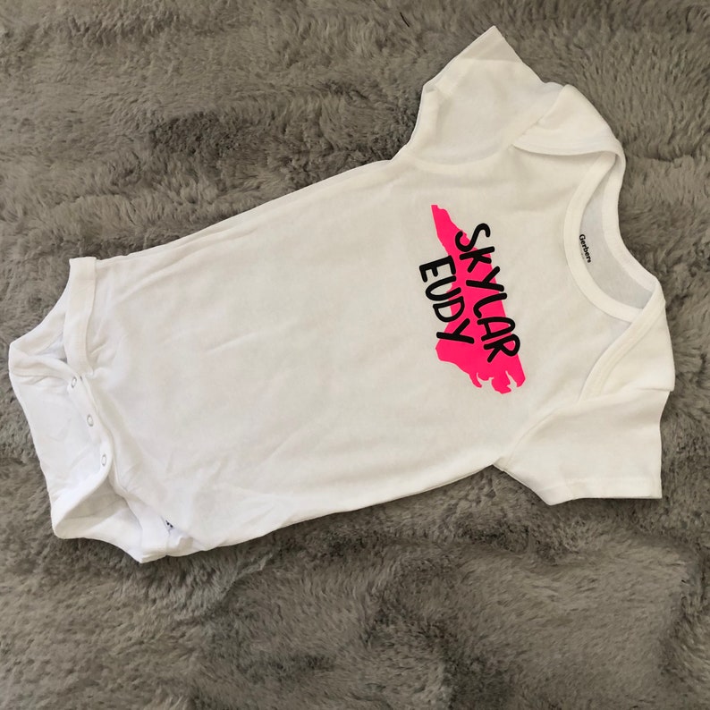 May include: White baby onesie with a pink outline of the state of Tennessee and the text "Skylar Eudy" in black.
