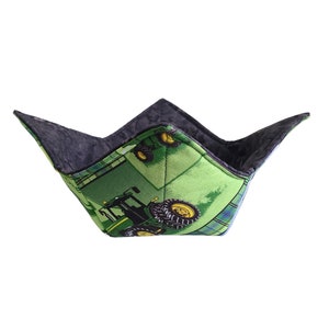 May include: A green and black bowl cozy with a tractor design. The exterior features a green background with a tractor and yellow wheels. The interior is a dark gray fabric. The bowl cozy is designed to hold a bowl of hot food.