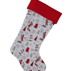May include: A Christmas stocking with a red cuff and a white body patterned with skiing snowmen, red trees, and gray snowflakes. The stocking has a loop for hanging.