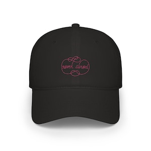 May include: A black baseball cap featuring an embroidered design in pink thread on the front panel. The embroidery consists of a decorative, swirling border surrounding the lowercase text "never cursed" in a cursive, script-style font.