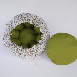 以下が含まれることがあります： 緑色の内装と、赤と緑の葉模様の白い外装の丸いペットベッド。 別売りの、お揃いの緑色のクッションがベッドの横にあります。 ベッドには巾着の留め具が付いています。