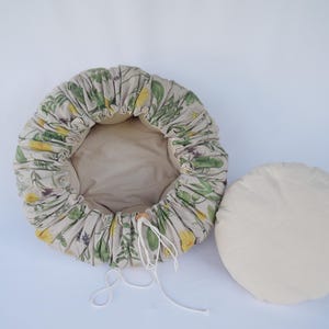 Peut inclure: Un lit pour animaux de compagnie beige avec un motif floral vert et jaune, à côté d'un coussin rond crème séparé. Le lit a un bord froncé et une fermeture à cordon. Le coussin mesure environ 30 cm de diamètre.
