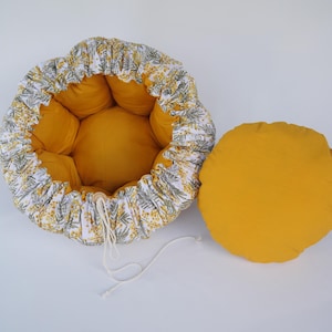 以下が含まれることがあります： 花柄模様の外側と無地の黄色の内側を備えたペットベッド。別の丸い黄色のクッションがベッドの横にあります。ベッドの外側は、黄色と緑の花柄のアクセントが付いた白い背景が特徴です。