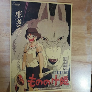 May include: A vintage style poster for the animated film "Princess Mononoke" featuring a young woman with red markings on her face standing next to a large white wolf. The poster is in Japanese and features the title "もののけ姫" (Mononoke Hime) in large red letters.