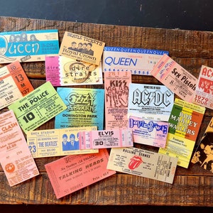 K&ouml;nnte beinhalten: Eine Sammlung von Vintage-Konzertkarten mit Bands wie Queen, The Who, The Police, The Clash, The Beatles, Ozzy Osbourne, KISS, AC/DC, Sex Pistols, Bob Marley, The Velvet Underground, Talking Heads und Elvis Presley. Die Tickets sind auf einer Holzoberfl&auml;che angeordnet.