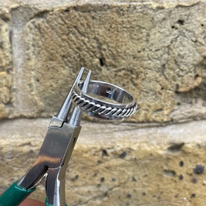 May include: A silver ring with a braided rope design around the outer edge is held by pliers with green handles. The ring is set against a textured stone background.