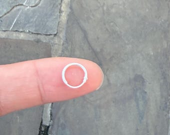 May include: A small, silver-colored hoop earring. The earring is a simple, circular design. The earring is held by a finger against a stone background.