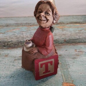 May include: A ceramic figurine of a woman wearing a party hat and holding a cup. She is sitting on a red block with the letter "T" on it.