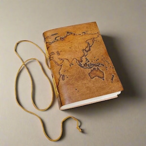 May include: A tan leather-bound journal featuring a dark brown engraved world map on the front cover. The journal is secured with a long, thin matching leather cord that wraps around the exterior, and it contains white paper pages visible at the edge.
