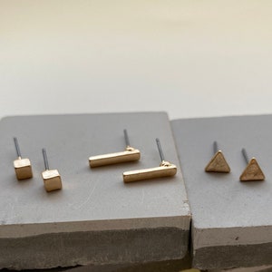 May include: A set of six gold-toned earrings with different geometric shapes: two squares, two rectangles, and two triangles. The earrings are displayed on two gray rectangular surfaces.