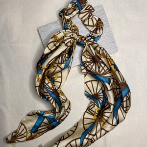 May include: A cream-colored hair scrunchie with a blue and brown pattern of repeating circles and chains. The scrunchie is tied in a bow.